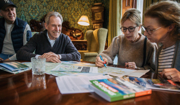 Drie mensen zitten rond een tafel met een kaart en bespreken de plannen voor hun Ierland reis. Een vrouw met een bril schrijft met een pen, terwijl een man lacht en een ander persoon luistert. Een reisgids is zichtbaar op de tafel. De kamer is warm verlicht met vintage decor.